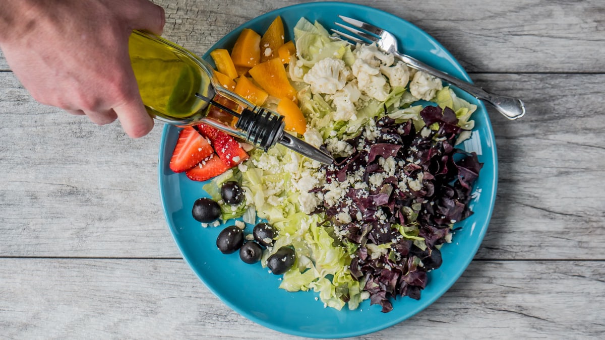 A vibrant vegetable salad with olives, feta, and strawberries being drizzled with olive oil.